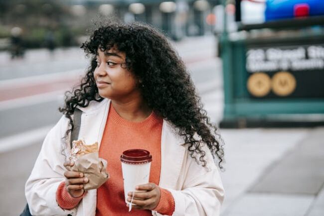 A woman holding a sandwich and a cup while standing on the street